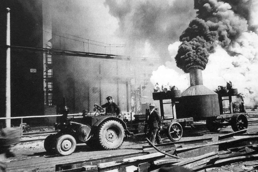 Traktor Lanz Bulldog jede po ulici chemického závodu a táhne speciálně upravený valník s kamny a koryty vytvářejícími umělou mlhu.